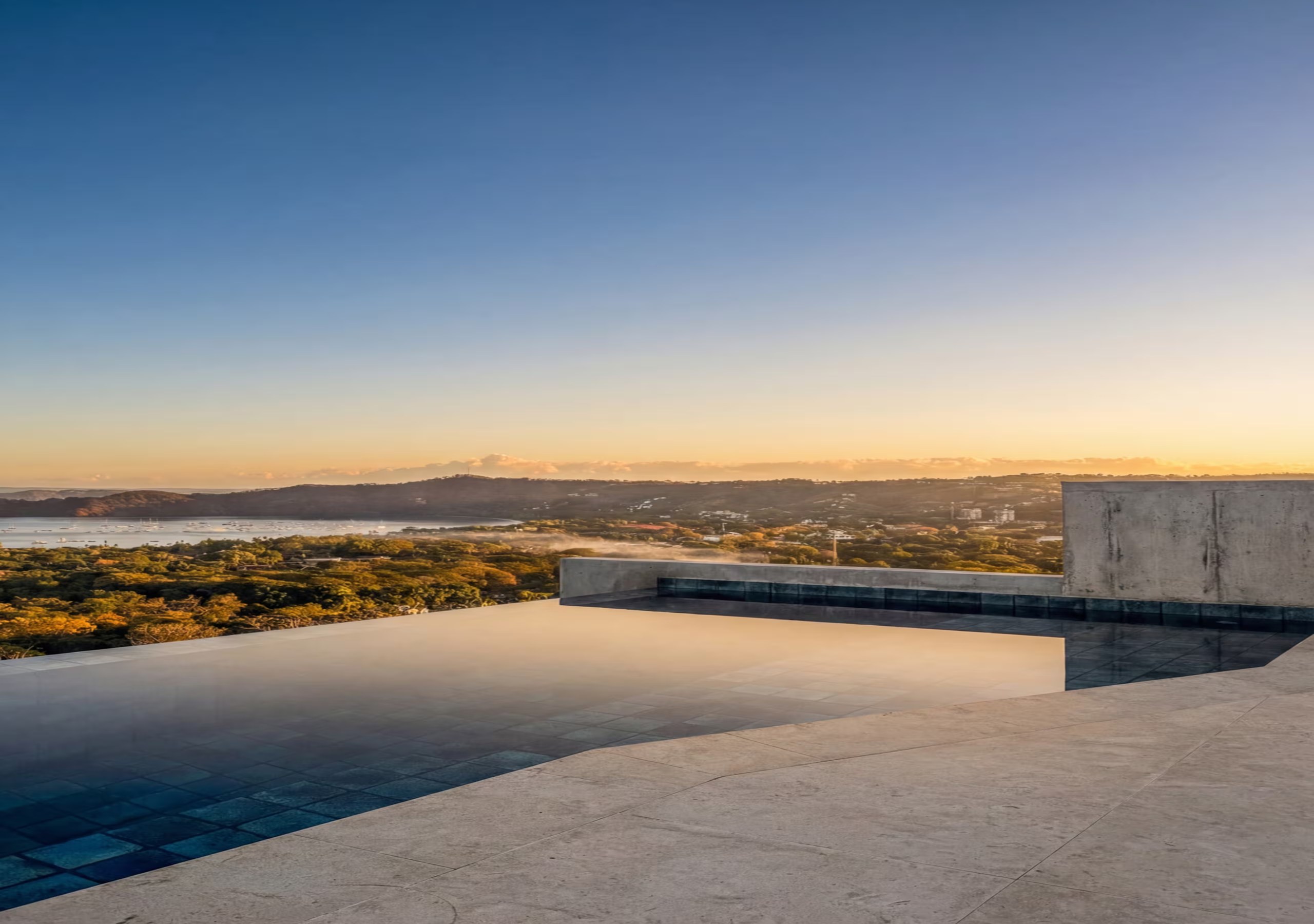 An infinity-pool terrace at golden hour: dark pool water at the left, pale limestone coping and deck across the foreground, a board-formed concrete parapet at the right, and a lit coastal plain and low hills under a blue-to-amber sky.